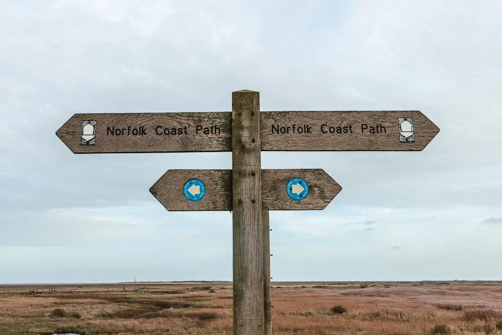 A wooden Norfolk Coast Path sign pointing left and right, with a backdrop of marshland, on the walk between Hunstanton and Burnham Overy Staithe.