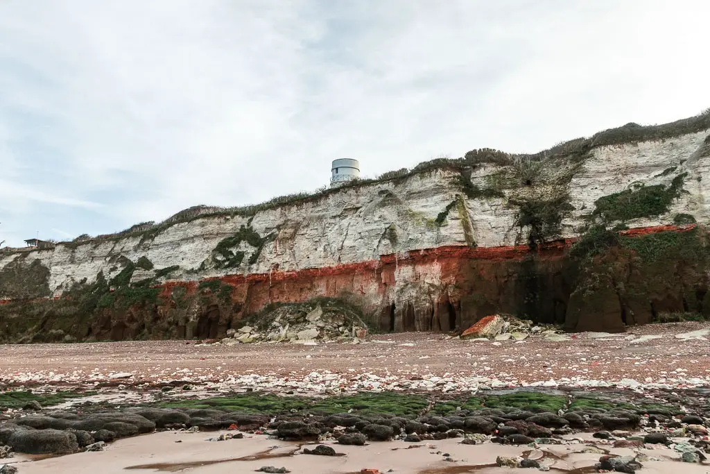 Looking straight towards the red and white stripped Hunstanton Cliffs, with the sandy rocky beach below, at the start of the walk towards Burnham Overy Staithe. There is the top of a lighthouse visible behind the cliffs.