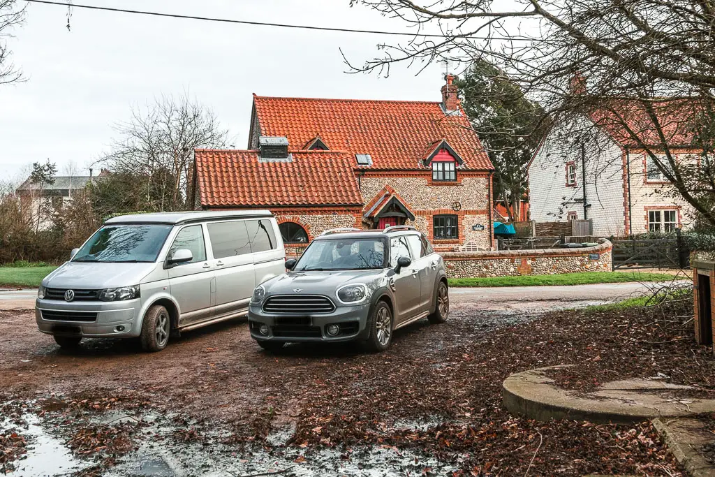 Two parked cars and a road behind them, with a stone walled house on the other side.