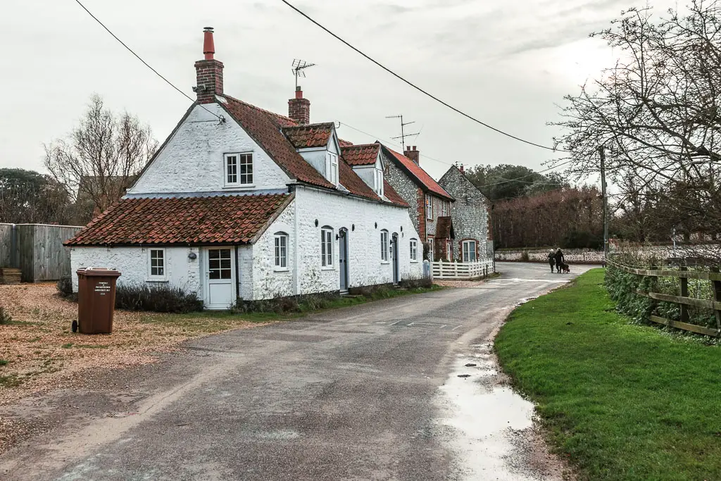 A residential road, with a strip of grass on the right, and white walled house on the left.