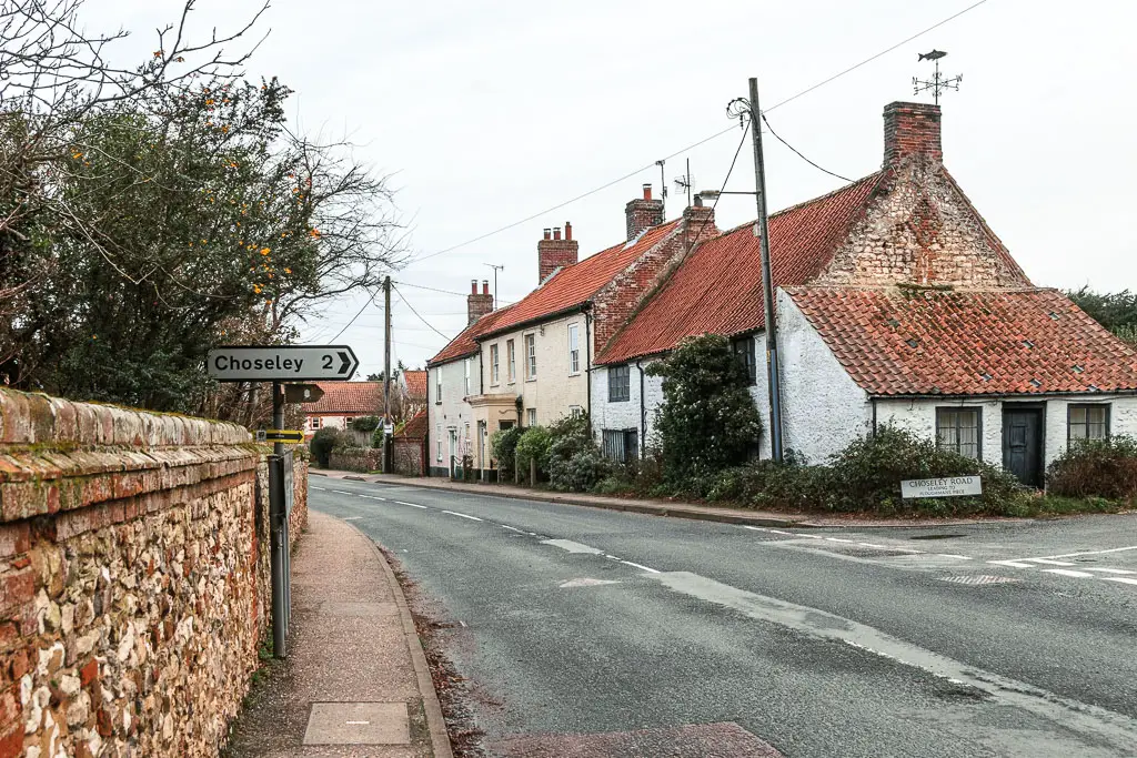 A road leading ahead and curving left. There is a stone wall on the left side of the road, and houses on the right. There is a sign on the left side of the road pointing right for Cholseley.