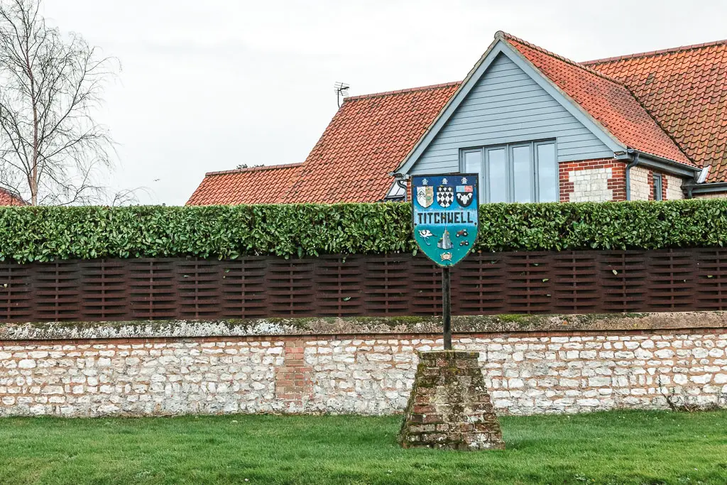 A titchwell coat of arms sign sitting on a pole on a strip of grass, with a wall behind it, and the rooftop of a house on the other side of the wall. The wall is stone art the bottom, then wooden fence above, and topped with a neatly cut green hedge.