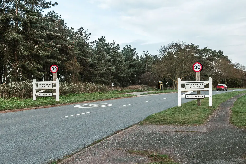 The road leading past signs which say 'Brancaster Staithe, slow down'. There is a red car driving along the road ahead.