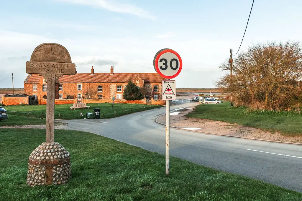 A medieval Brancaster Staithe sign on the grass, next to a 30 speed limit road sign. the road is on the right, leading towards terraced houses ahead.