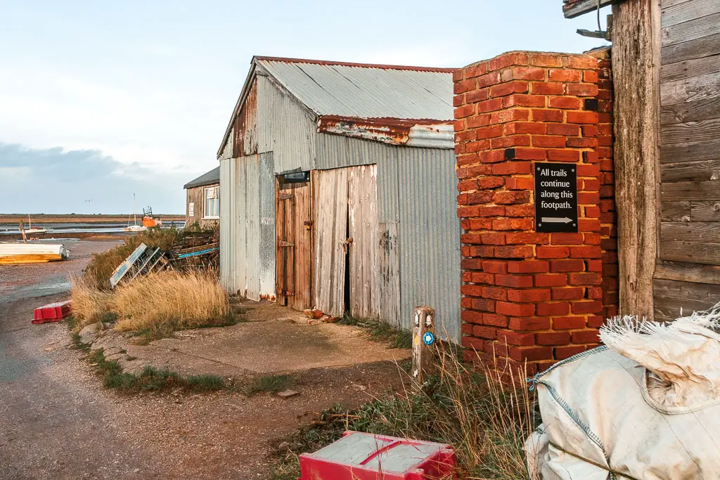 The road on the left and a brick pillar on the right next to a rusty shed. There is a black sign on the brick pillar pointing right for the footpath trails.