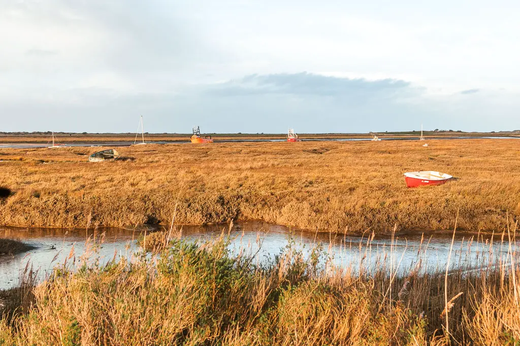 Looking across the orange/brown marshes, with a stream running across, part way through the walk from Hunstanton to Burnham Overy Staithe. There are a few boats strewn across the marshes.