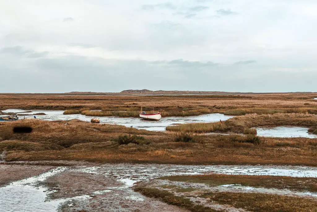 Looking across the marshland with strips of water, and a small boat resting in the river ahead, near the end of the walk from Hunstanton to Burnham Overy Staithe.