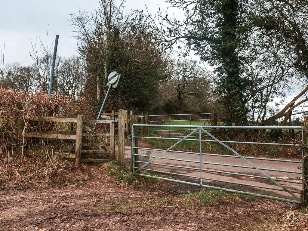 A metal gate and wooden gate on the end of a field, with a road on the other side.