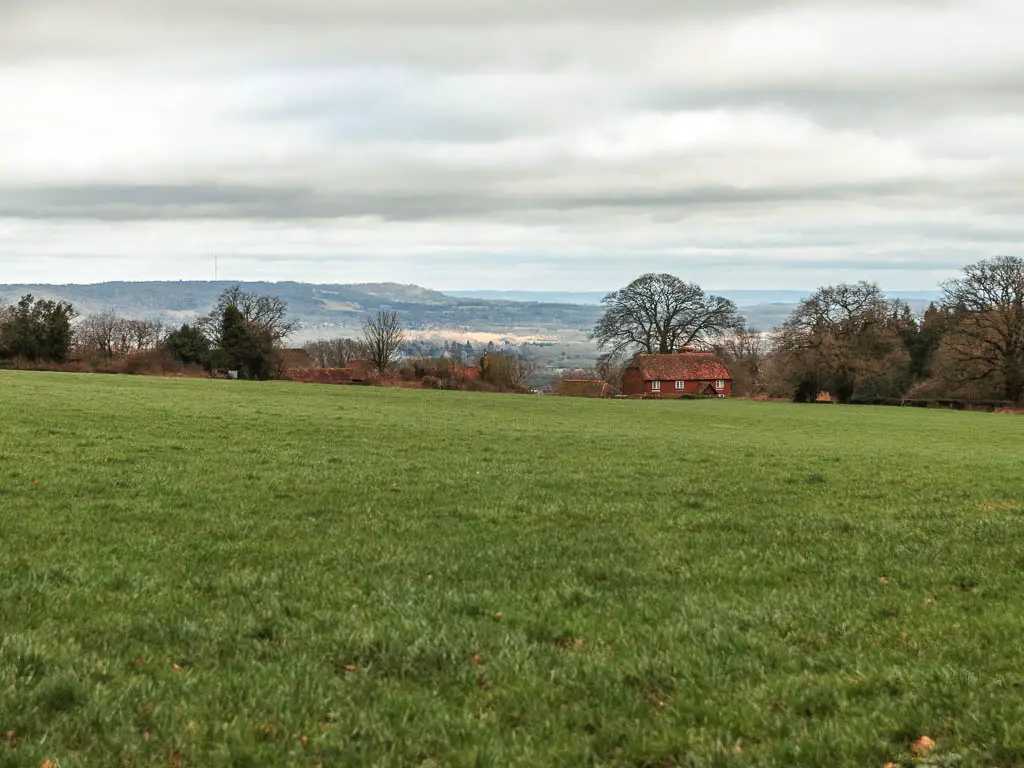 Looking across a large green grass field on the walk from Oxted to Otford. There is a cottage on the other side of the field, and hills in the distance.