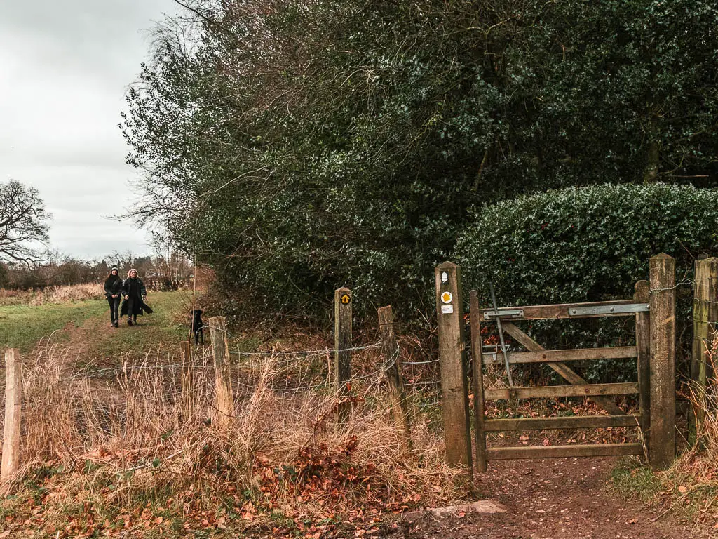 A wooden gate and wire fence on the edge of a field, with hedges and bushes to the right. There are two people walking in the field on the other side of the gate.