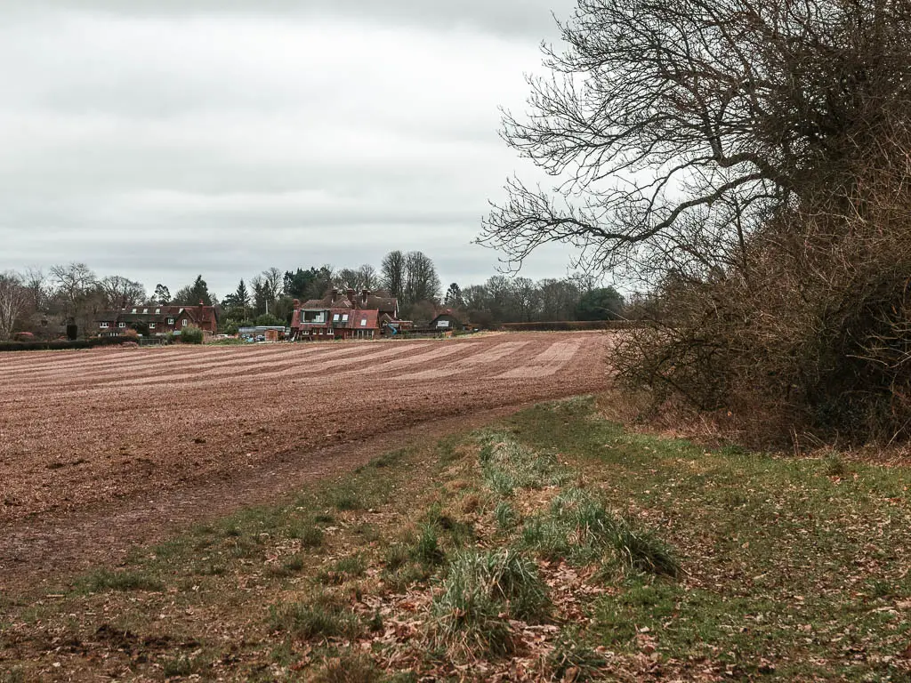 Looking across an empty crop field, to a farm house on the other side, partway through the walk between Oxted and Otford. There are leafless bushes and trees on the right side of the field.