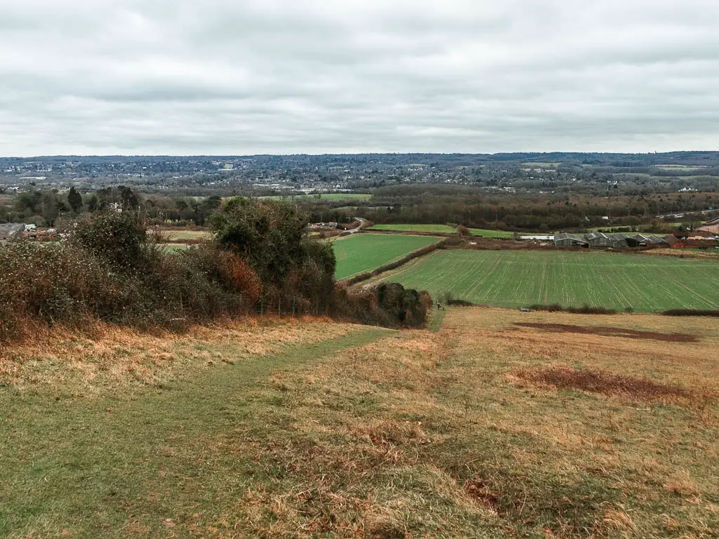 Looking down the hill field, to a view of patchwork fields below. The iOS a grass Traill running down the hill, and bushes lining the left side.