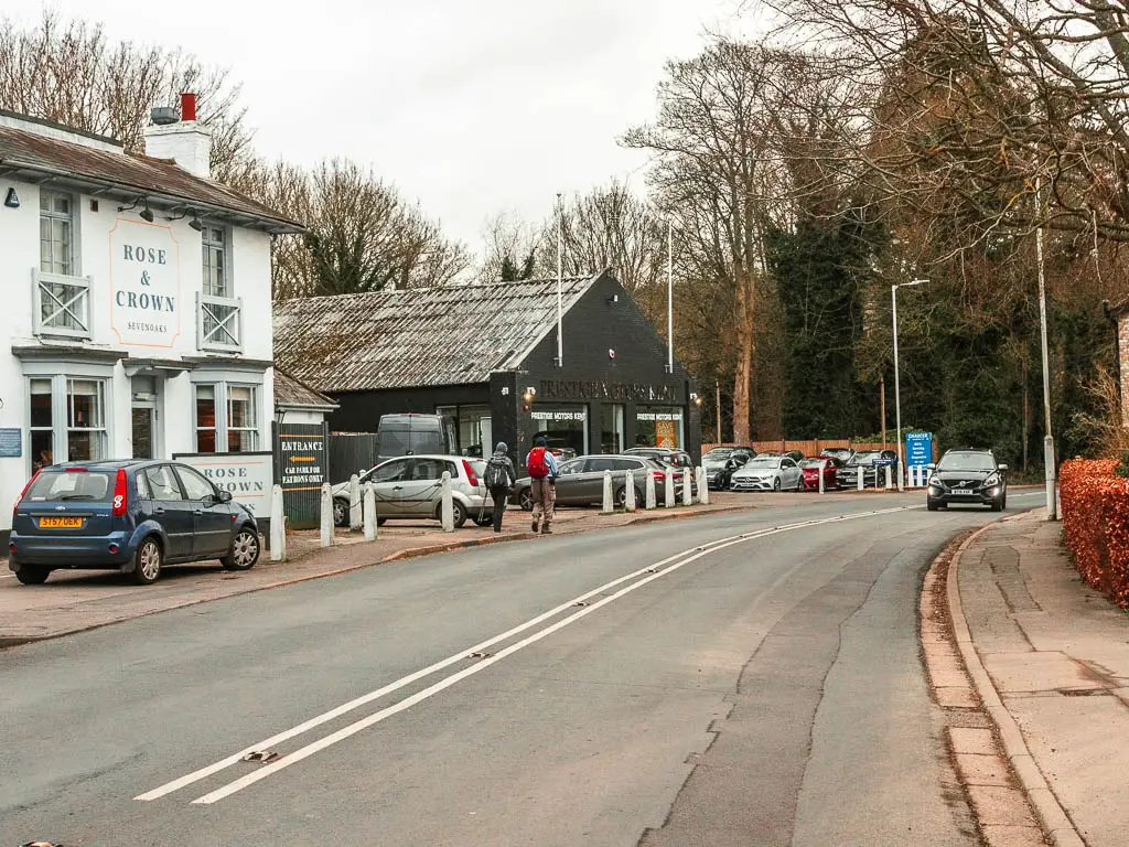 A road with the white walled rose and crown pub on the left side. There is a black car driving on the road, and cars parked on the pavement on the left side. There are two people walking on the left side pavement.