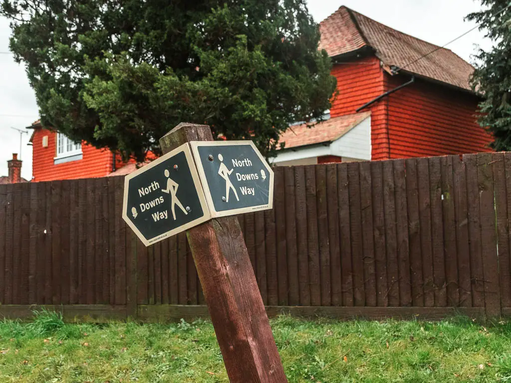 A leaning North Downs way sign pointing left and right, with a brown wall behind it, and a red bricked house on the other side.