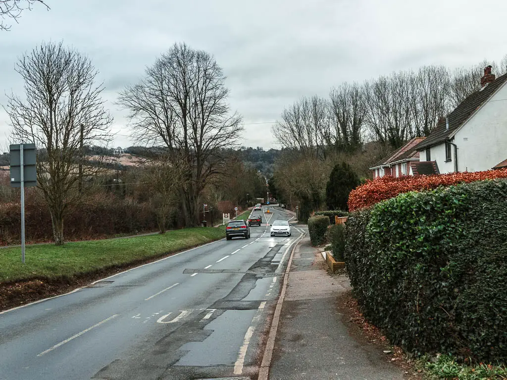 Looking along the main road with cars driving along it. There are hedges on the right and the walls of a white coloured house behind the hedges.