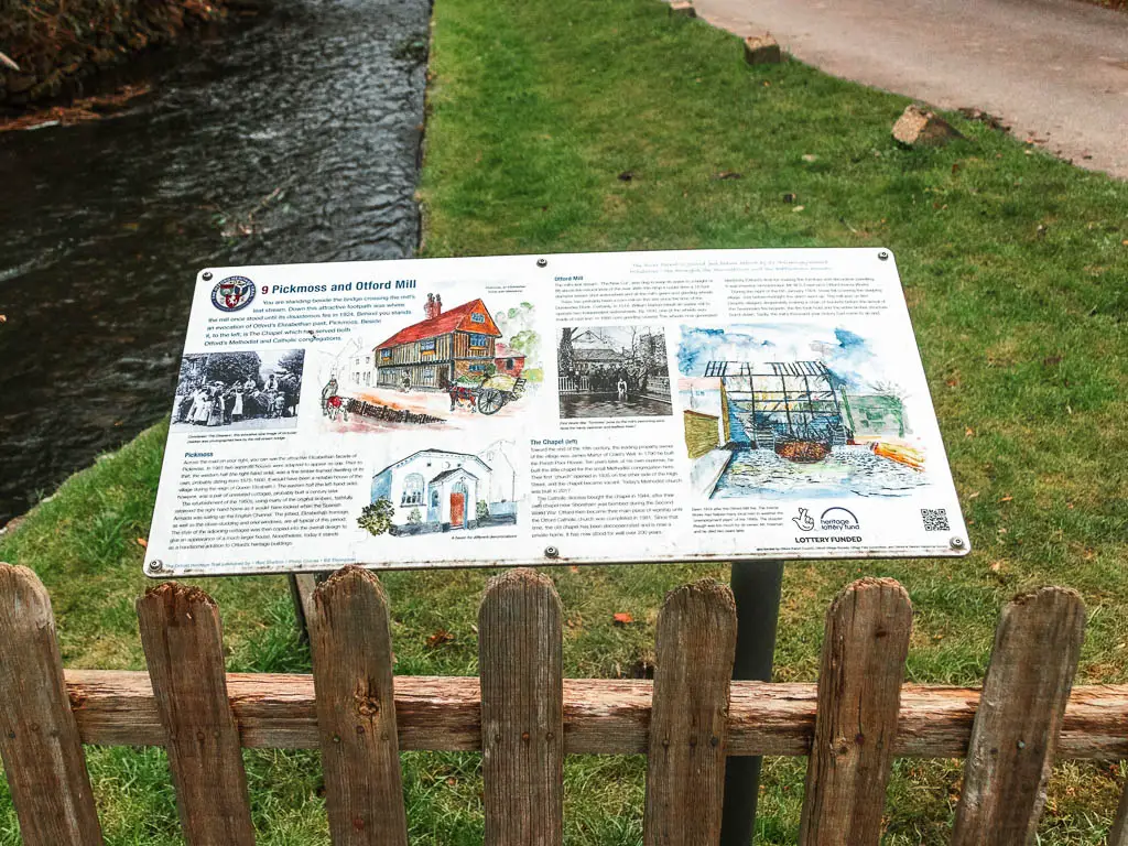 An information board with drawings sitting behind a wooden fence eon the grass. There is a stream of water to the left.