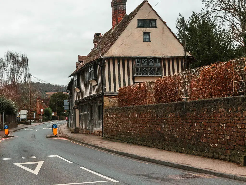 An old Tudor building on the road side of the road, with a stone wall leading to it.