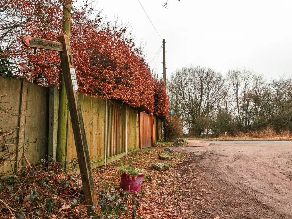 A wooden trail signpost on the left, next to a wooden wall with a red leafed bush coking over the top, The road is ahead to the right.