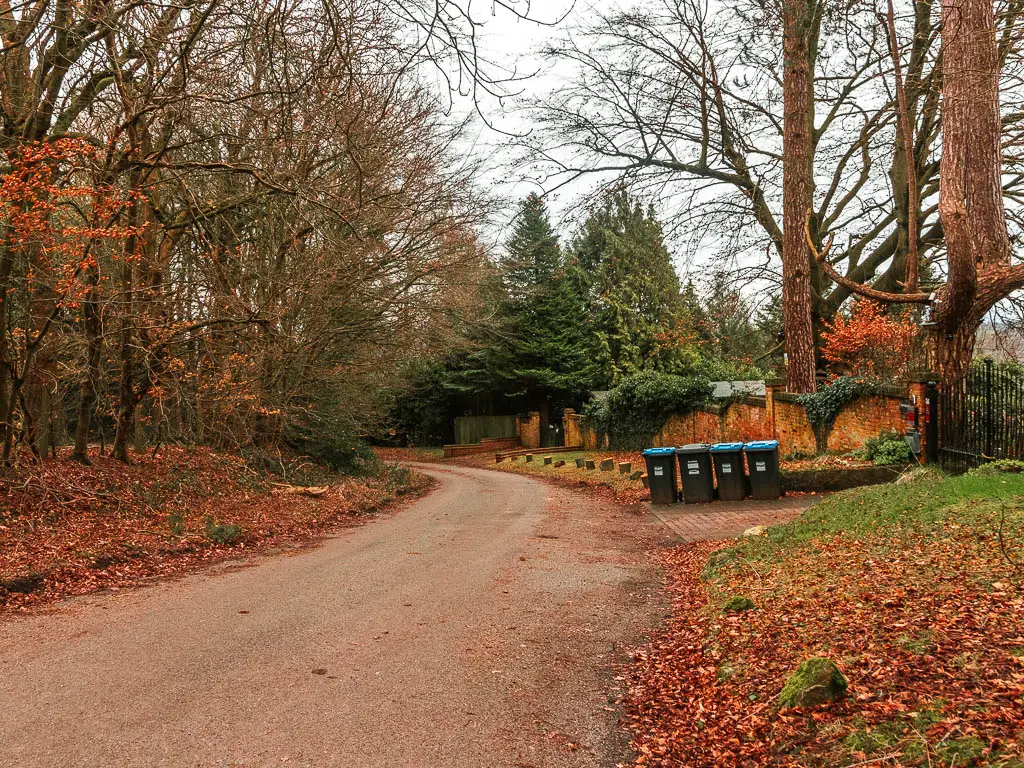 A road curving to the left, with trees to the left side, and a wall and gate entrance to a home on the right. There is a row of bins ahead on the right.