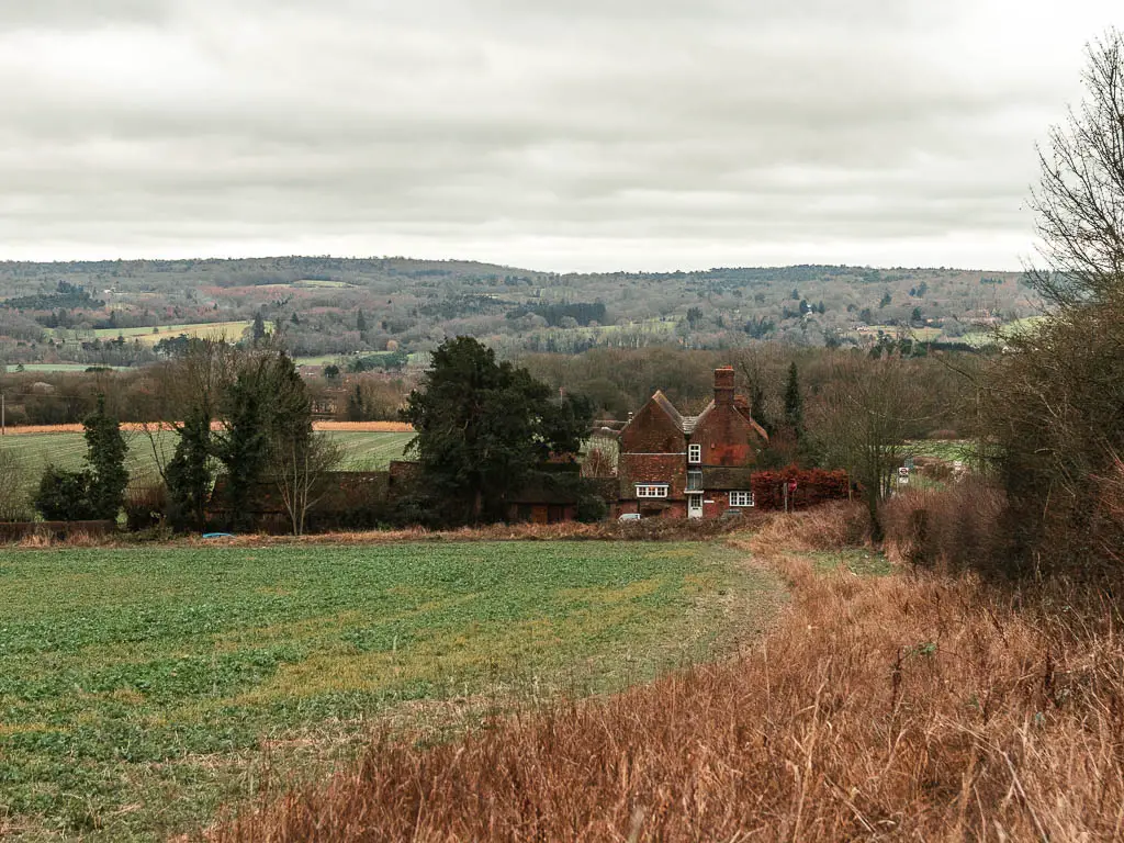 Looking along the edge of a green grass field, towards a cottage on the corner, when walking between Oxted and Otford. There are hills reaching far into the distance. 