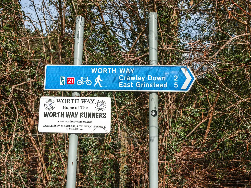 A blue sign infant of trees and bushes, pointing right to walk along the Worth Way towards east grinstead and Crawley down. 