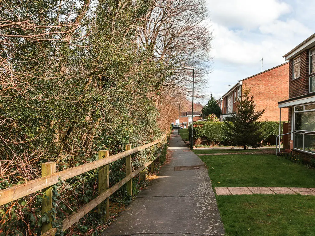 A path with a wooden fence and bushes on the left, and houses with small green front lawns on the right.