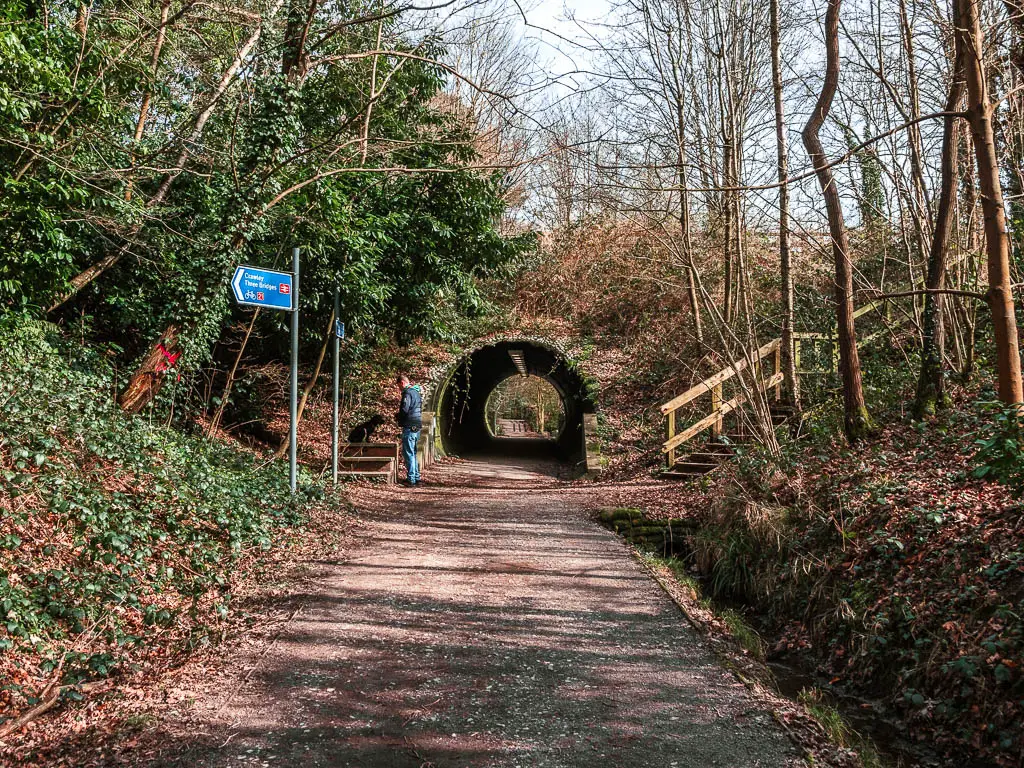 A wide path leading towards a bridge tunnel. The bath is lined with dirt banks and trees. There is a man standing next to a blue sign ahead.