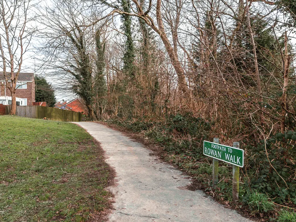 A path leading ahead, with grass to the left and straggly leafless trees on the right. there is a green road sign on the right saying 'footpath to rowan walk'.