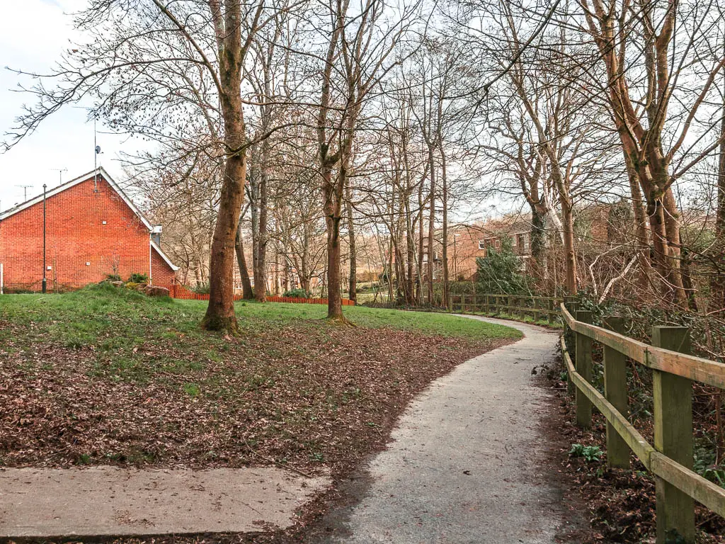 A path winding ahead, with a green on the left and wooden fence on the right. There are a couple of trees on the tree, and a red brick walled house past the green.