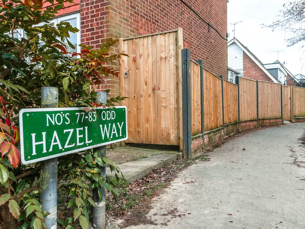 A green road sign on the left side of the path saying 'Hazel road' . There is a wooden fence lining the path on the left, with houses on the other side.