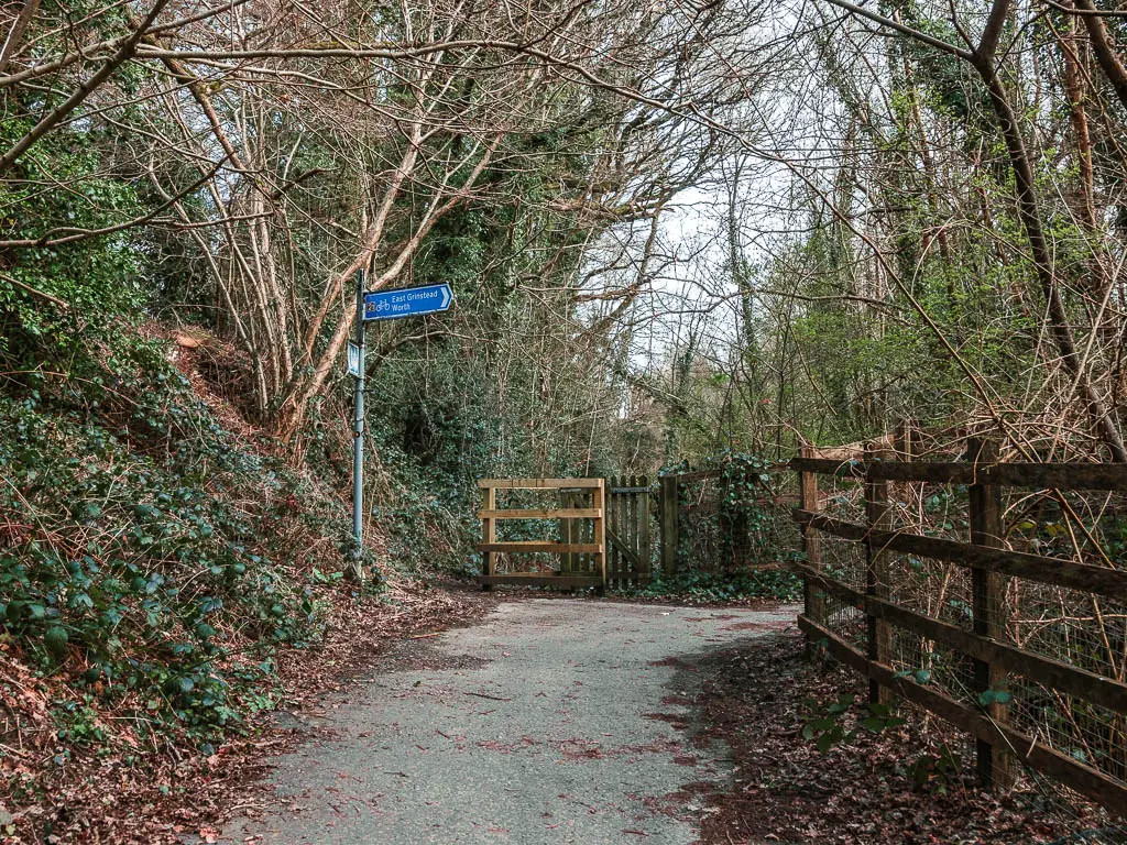 A path leading to a gate, surrounded by trees. There is a wooden fence on the right, and bush covered bank on the left. There is a blue sigh ahead, pointing right.