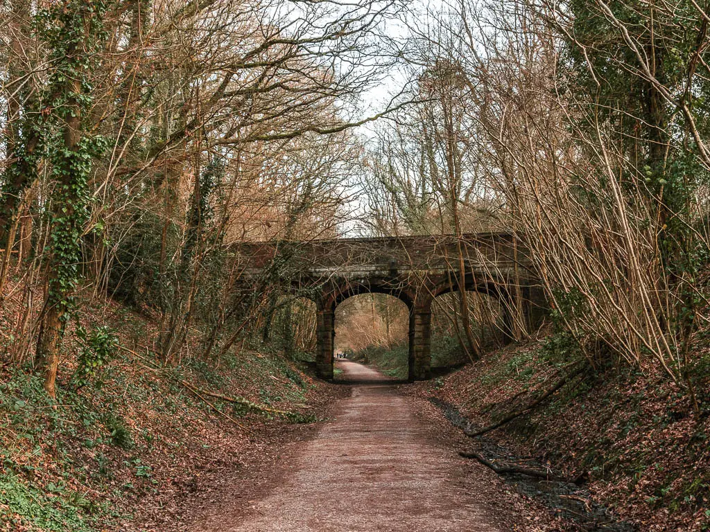 A wide path leading towards an archway bridge, near the end of the walk along the worth way. The path is lined with steep dirt banks and straggly tree branches.