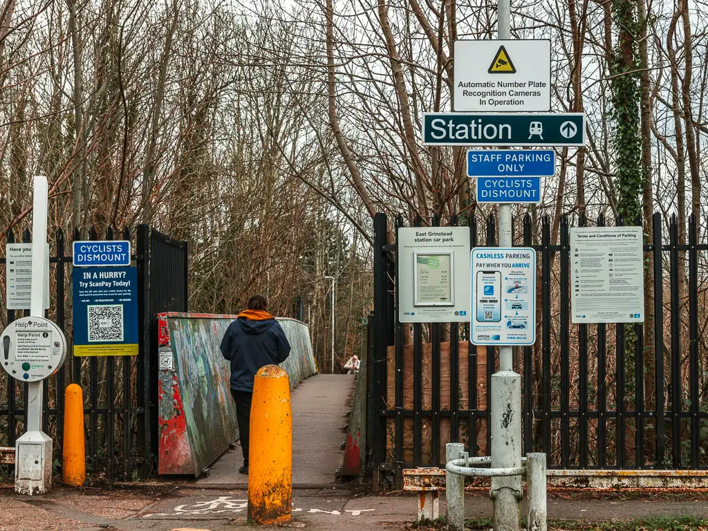 A path leading past a metal fence with a sign in a lamppost saying 'station'. There is a person walking g on the path. There is an orange pillar in front of the path.