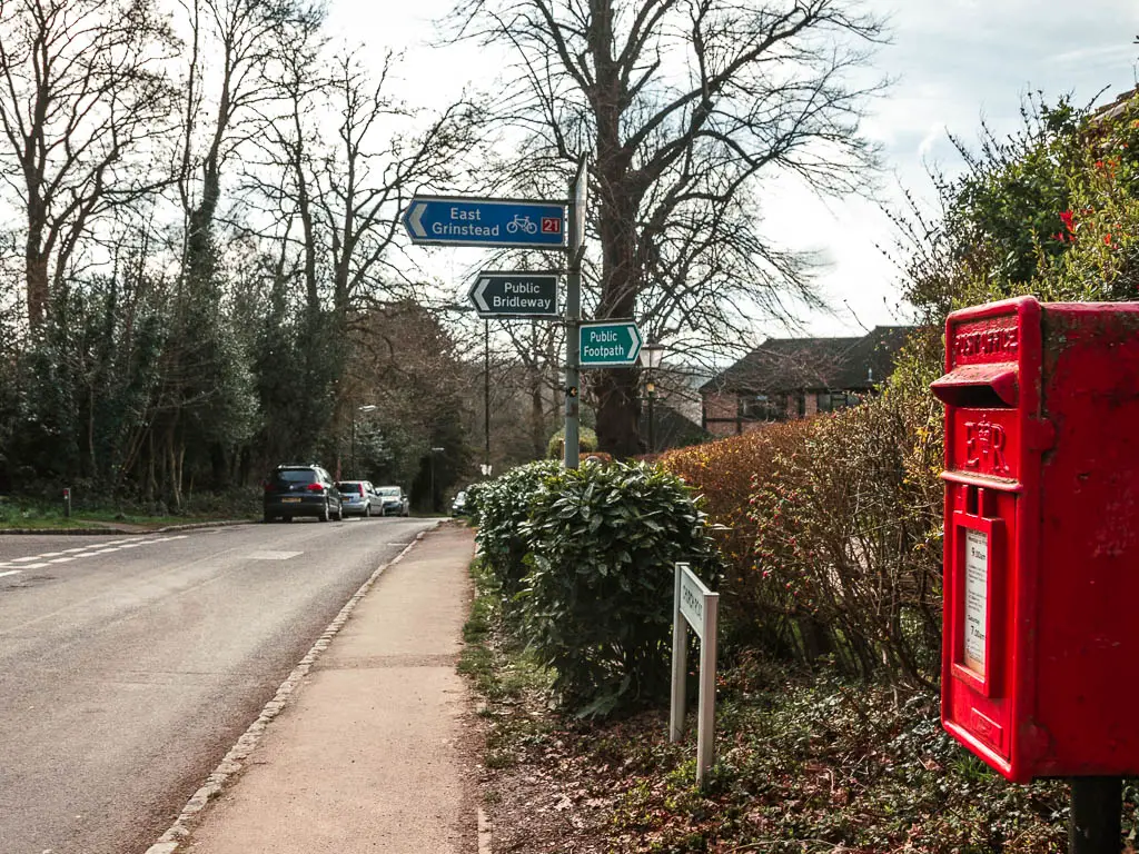 A pavement with a road on the left. There is a red post box on the right, and signposts ahead.