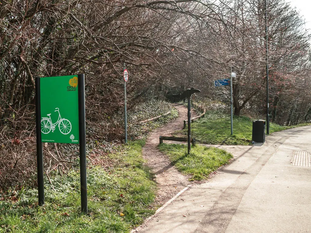 A dirt trail leading off the pavement into the leafless woodland. There is a green sign on the left.