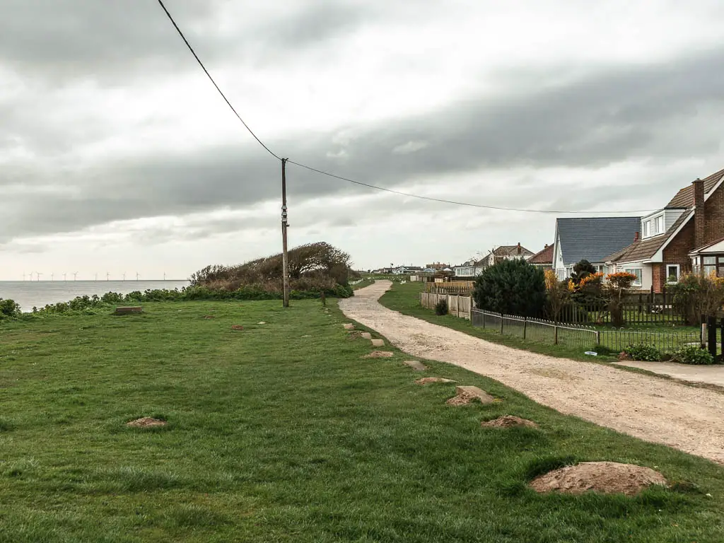 A large green with a gravel type road on the right, with houses lining the right side. There is a telephone pylon wire running across the frame.