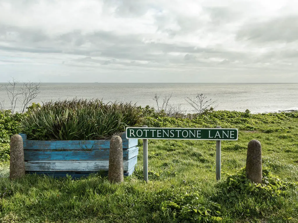 A road sign saying 'Rottenstone Lane' on the green, with the sea in the distance. 