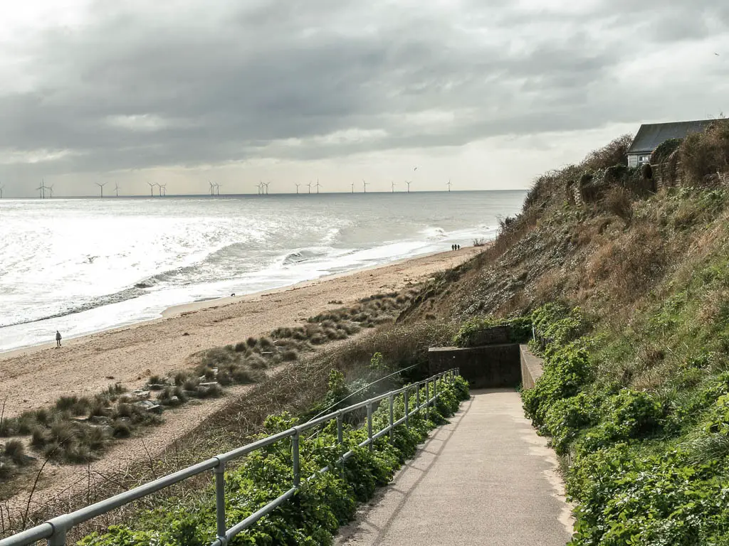 A concrete walking way leading down to the beach. The sandy beach is to the left and grass covered cliffs to the right. There is a person walking on the beach with the sea to the left. The sea is glistening white form the sun.