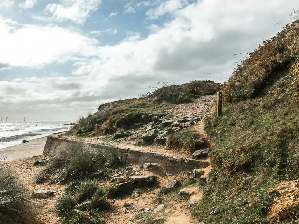 The end of the beach with the sea wall and path leading up it to the top of the grass covered cliffside.