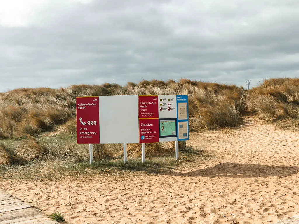 An information board on the sand in Caister on Sea. There is tall yellow and green grass behind it.