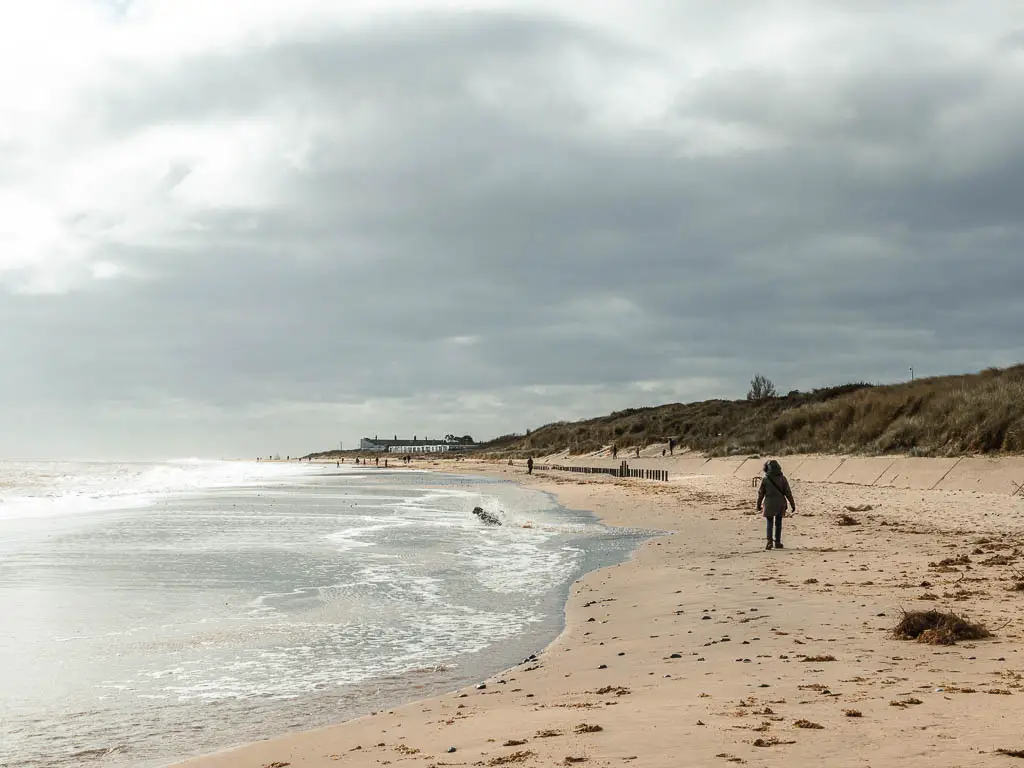 Where the sea meets the sand on the walk from Hemsby to Great Yarmouth. The sand is on the right and a cream colour and the sea is on the left glistening white from the sun. There is a person walking along the beach wearing all black.