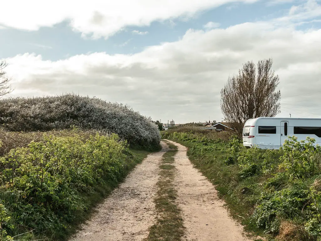 A track type trail lined with green bushes. There is the back of a white caravan on the right side.