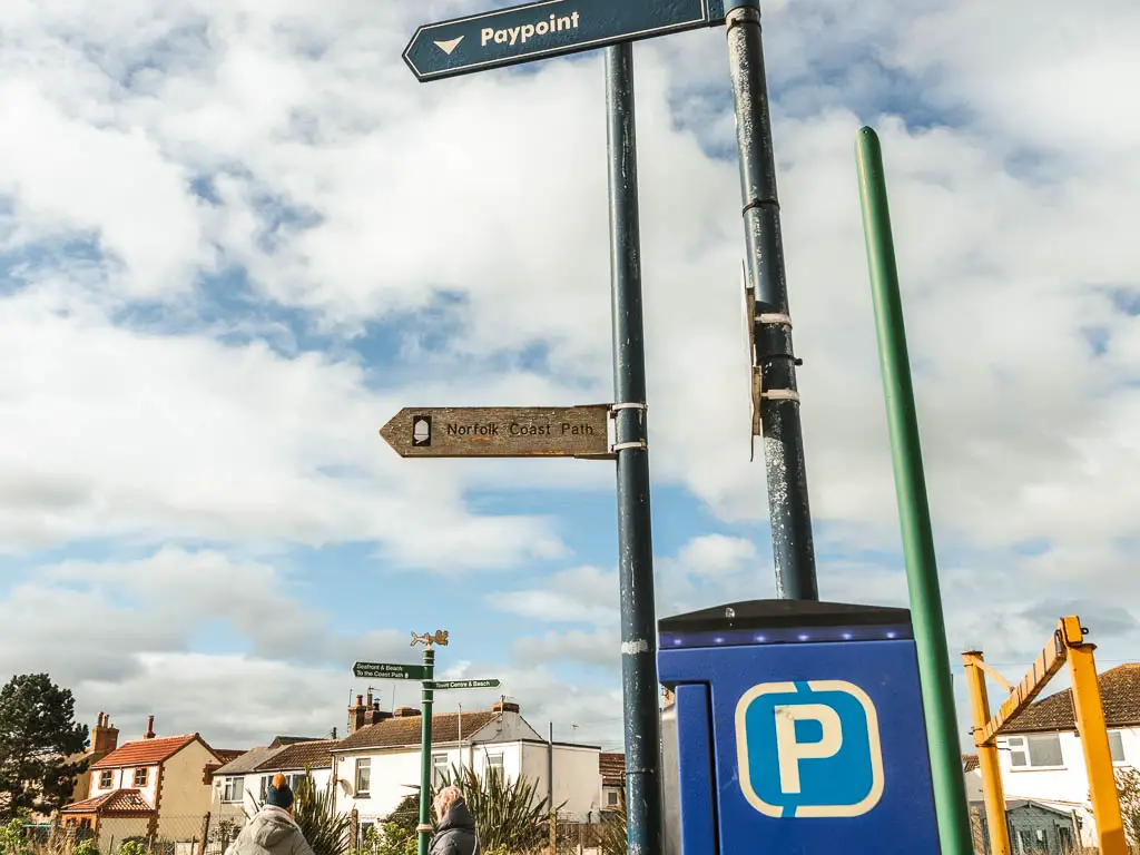 A coast path sign on a lamppost, next to a blue car parking payment machine. There are houses further back in the background.