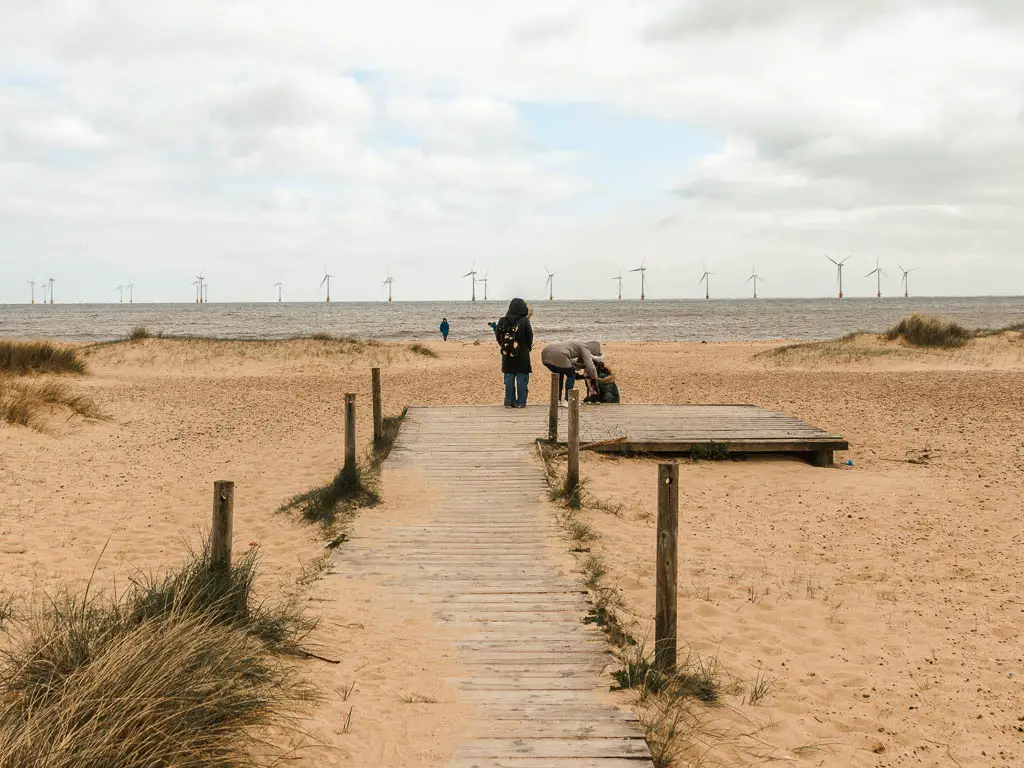 A wooden walking leading to the sandy beach on the walk between Hemsby and Great Yarmouth. There are people standing at the end of the walkway. The sea is ahead in the distance.