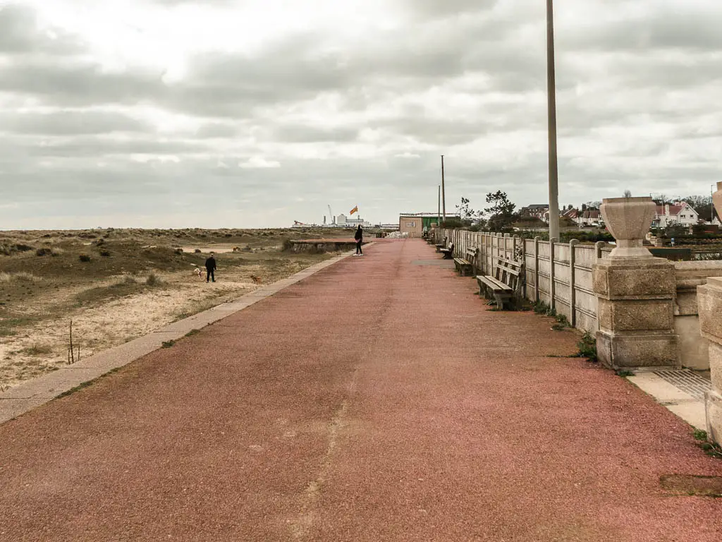 A red wide promenade with a wall and benches on the right and grass to the left.