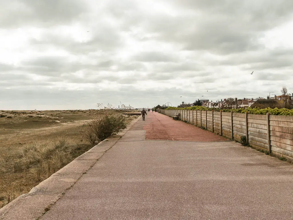 A wide promenade leading straight ahead to Great Yarmouth in the distance. There is a wooden wall on the right, and grass below to the left. There is a person walking ahead. 