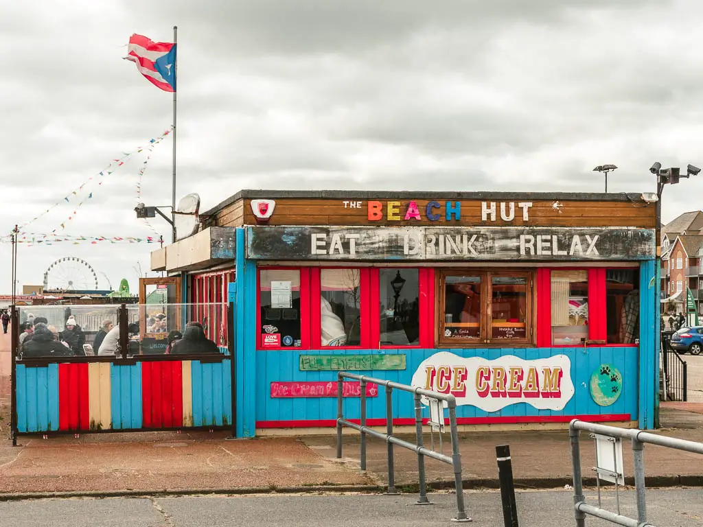 A colourful blue and red beach hut selling food. There are people sat outside on the left eating.