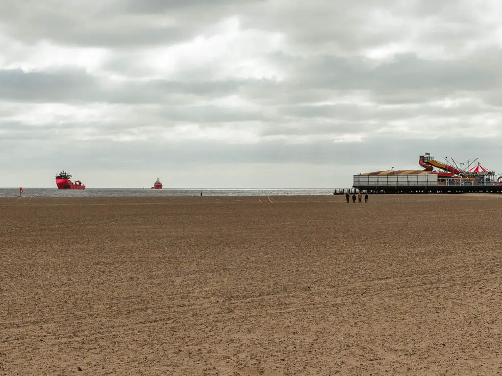 A very big almost empty sandy beach in Great Yarmouth with just 4 people walking on it in the distance. There are two big red cargo ships in the sea and part of the promenade is visible far away on the right.