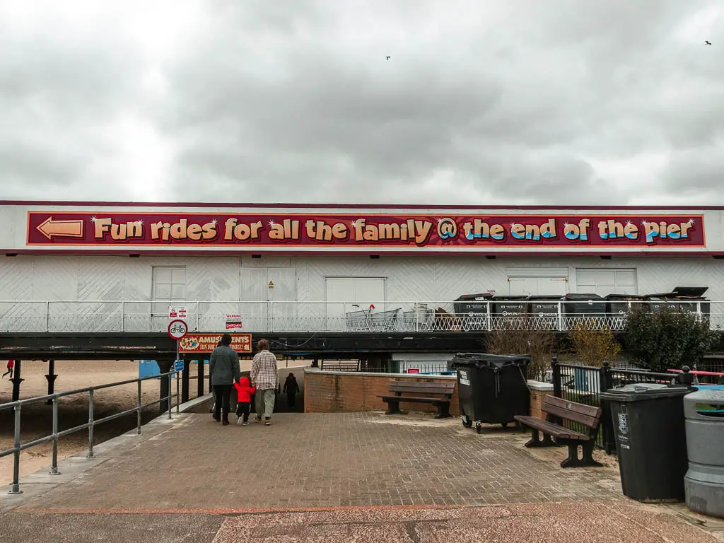 A wide walkway in Great Yarmouth with the promenade across the pier ahead, and a big sign saying 'fun rides for all the family at the end of the pier'. There are two adults and a child walking under the pier. 