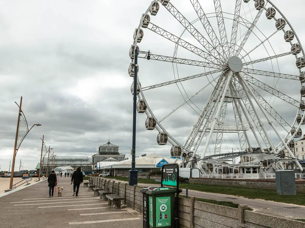 A big ferris wheel to the right, with the promenade to the left in Great Yarmouth. There are two people walking a dog on the promenade.